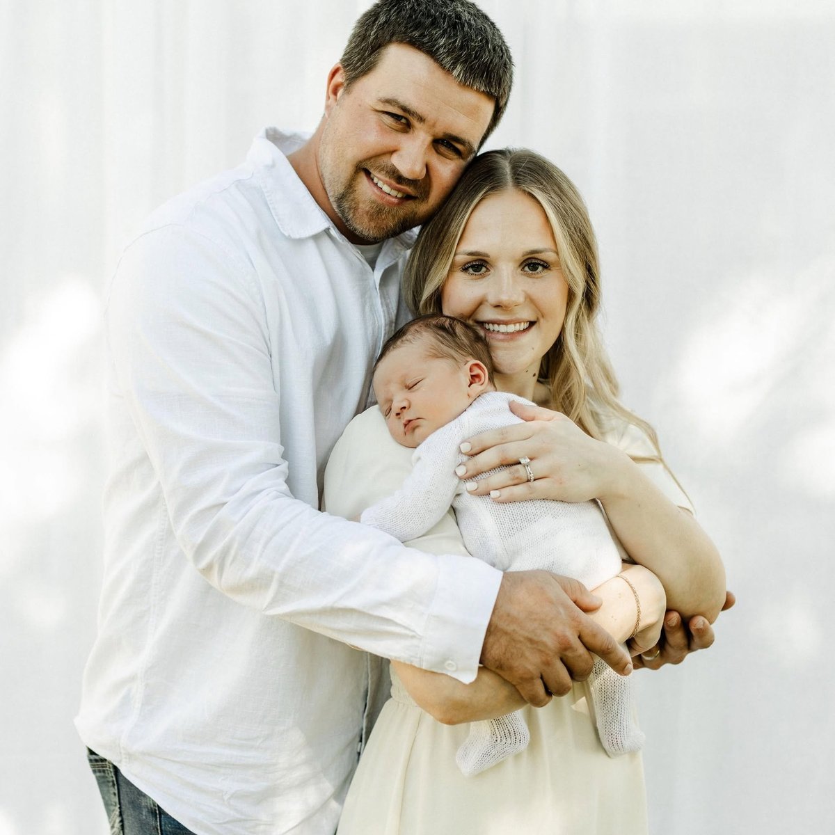 Cody and Kaitlyn smiling together while holding their newborn son wrapped in white